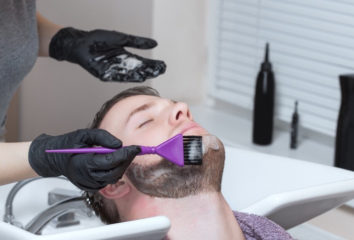 The barber paints the beard and the mustache of the young man in the barbershop.