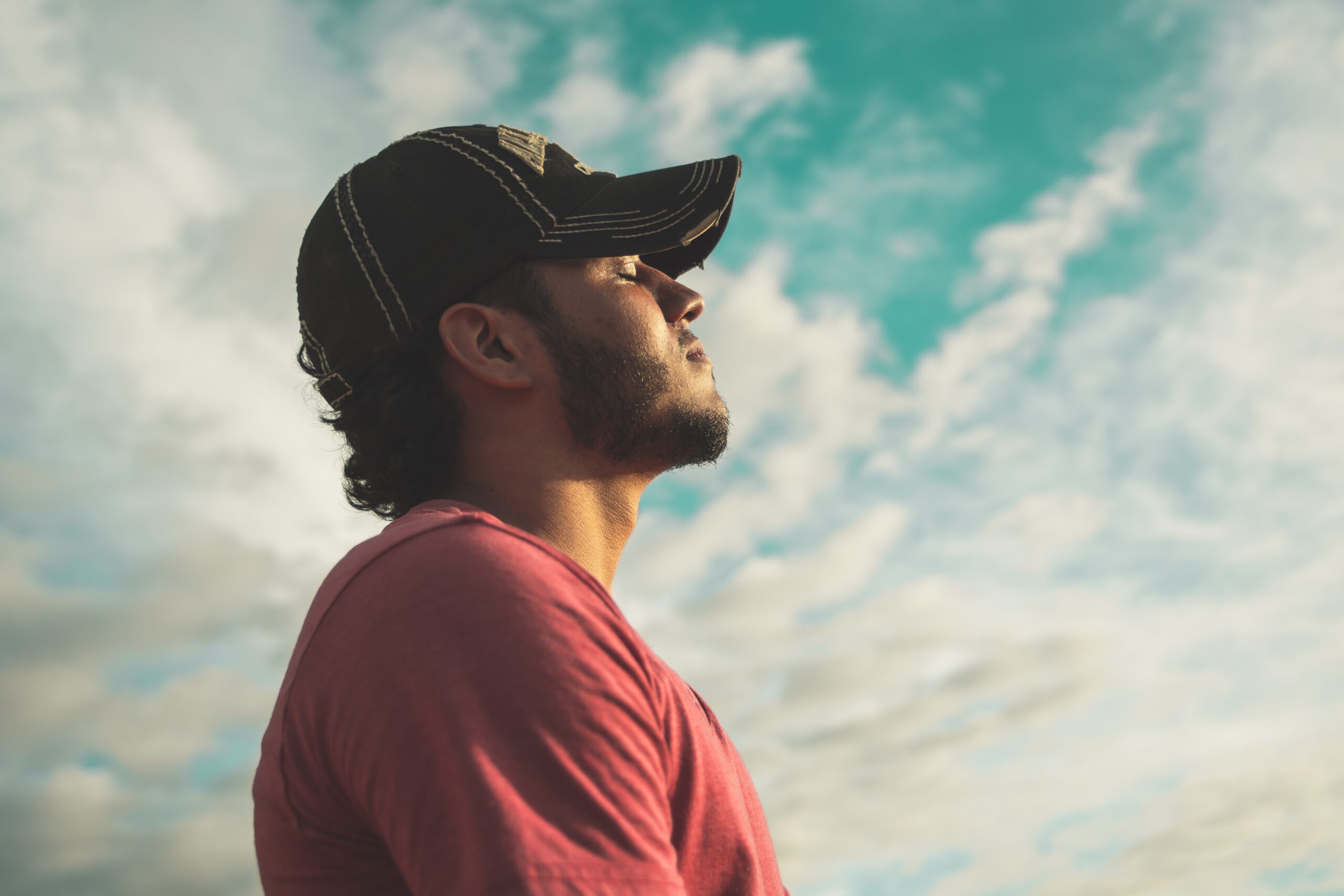 man wearing a black cap with eyes closed under a cloudy sky