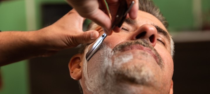 Barber shaves a client's cheek full of shaving foam with a razor in a barber shop.