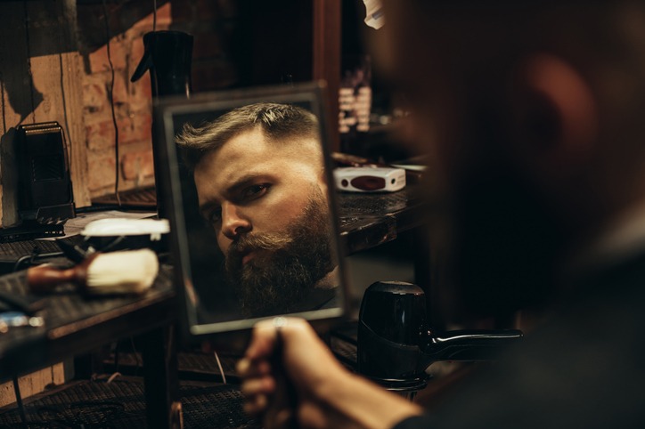 Young bearded man looking herself in the mirror while sitting in chair at a barbershop